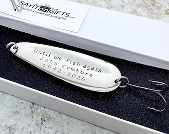 May include: A silver fishing lure with the inscription "Until we fish again John Cembura 1942-2026" is presented in a white gift box. The lure has a hook and is a memorial gift.