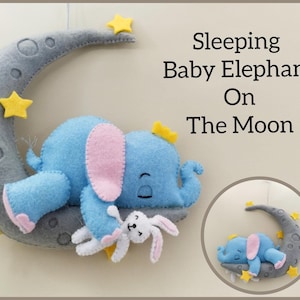 Peut inclure: Mobile pour bébé en feutrine représentant un éléphant bleu endormi avec des oreilles roses et une couronne jaune, reposant sur un croissant de lune gris. La lune est ornée d'étoiles jaunes et du texte "Sleeping Baby Elephant On The Moon". Un petit lapin blanc est également présent.