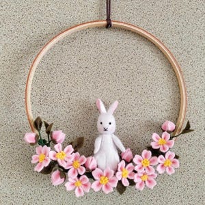 May include: A felt cherry blossom wreath with a bunny. The wreath has a wooden hoop decorated with pink felt flowers and green leaves. A white bunny is in the centre. The text "Felt Cherry Blossom Wreath with Bunny" is at the top.