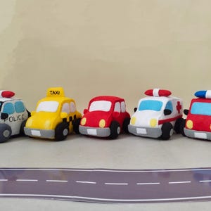 Puede incluir: Colección de cinco vehículos de juguete de fieltro de colores: un coche de policía, un taxi, un coche rojo, una ambulancia y un camión de bomberos. Cada coche tiene ruedas negras y detalles como luces y ventanas. Dispuestos sobre una carretera.