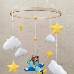 May include: A baby mobile featuring a wooden ring with hanging felt shapes. The mobile includes yellow stars, white clouds, and a blue airplane with a teddy bear pilot. The mobile is suspended by white strings.