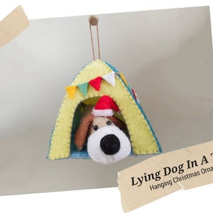 May include: A handmade felt Christmas ornament featuring a dog wearing a Santa hat inside a yellow tent with blue trim. The tent is decorated with colorful bunting. The text reads "Lying Dog In A Tent Hanging Christmas Ornament."
