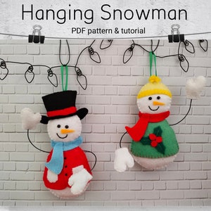 May include: Two felt snowman ornaments hanging from a string of lights against a white brick wall. One snowman is wearing a black top hat, red coat, blue scarf, and white mittens. The other snowman is wearing a yellow hat, green sweater, red scarf, and white mittens.
