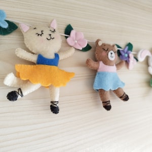 May include: A felt garland with a cat, bear, and bunny wearing tutus and holding flowers. The garland is strung with white thread and has green leaves and pink and purple flowers.