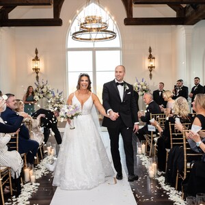 May include: A bride and groom walk down the aisle after their wedding ceremony, with guests throwing flower petals. The couple is smiling and holding hands. The ceremony is taking place in a white room with large windows and a chandelier.