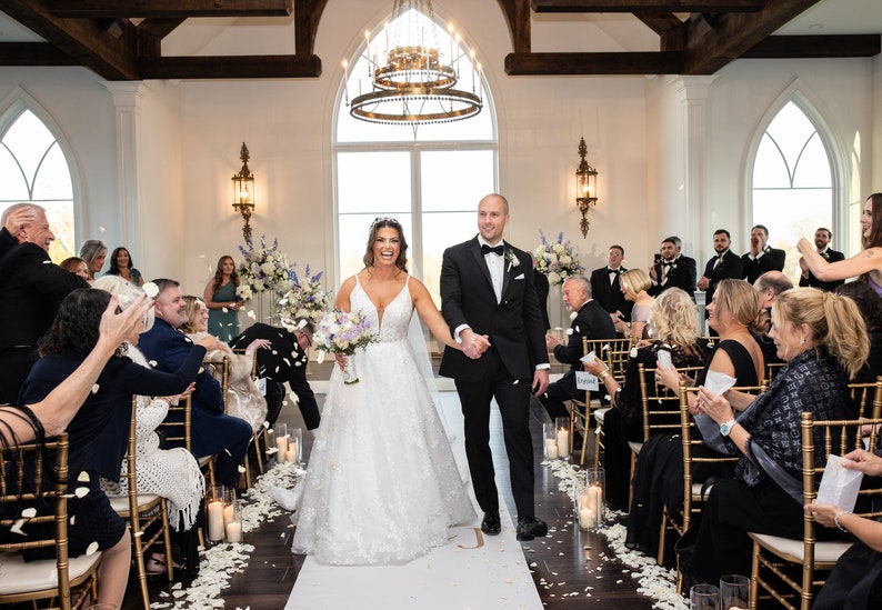 May include: A bride and groom walk down the aisle after their wedding ceremony, with guests throwing flower petals. The couple is smiling and holding hands. The ceremony is taking place in a white room with large windows and a chandelier.