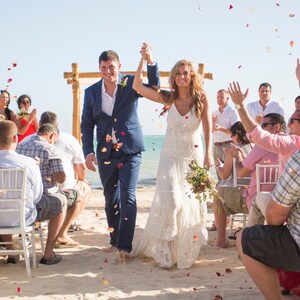 May include: A bride and groom walk down the aisle after their wedding ceremony on a beach. Guests throw flower petals as they exit the ceremony area.