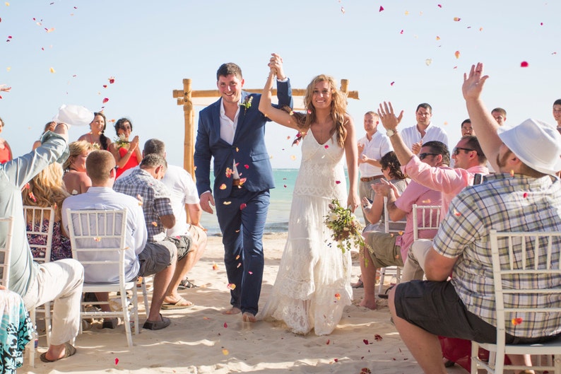 May include: A bride and groom walk down the aisle after their wedding ceremony on a beach. Guests throw flower petals as they exit the ceremony area.