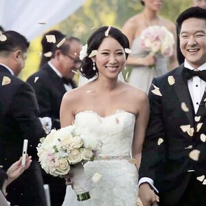 May include: A bride and groom walk down the aisle after their wedding ceremony, with flower petals being thrown in the air. The bride is wearing a white strapless wedding dress with a beaded belt, and the groom is wearing a black tuxedo with a black bow tie.