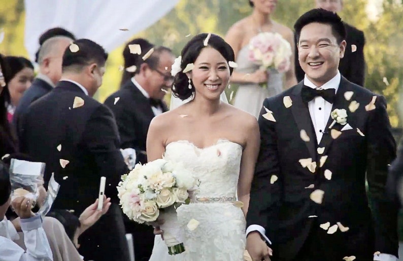 May include: A bride and groom walk down the aisle after their wedding ceremony, with flower petals being thrown in the air. The bride is wearing a white strapless wedding dress with a beaded belt, and the groom is wearing a black tuxedo with a black bow tie.