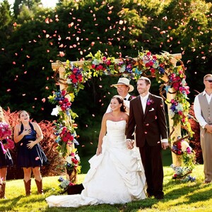 May include: A wedding ceremony taking place outdoors under a floral archway. The bride and groom are standing under the arch, surrounded by guests. The bride is wearing a white wedding dress and the groom is wearing a dark suit. Guests are throwing flower petals in the air.