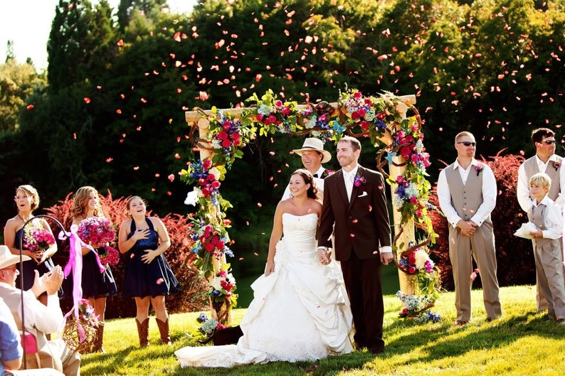 May include: A wedding ceremony taking place outdoors under a floral archway. The bride and groom are standing under the arch, surrounded by guests. The bride is wearing a white wedding dress and the groom is wearing a dark suit. Guests are throwing flower petals in the air.