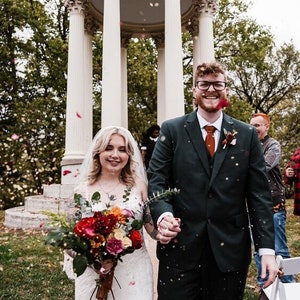 May include: A bride and groom walk down the aisle after their wedding ceremony, with red and white flower petals falling around them. The bride is wearing a white wedding dress with a veil and carrying a bouquet of red, orange, and yellow flowers. The groom is wearing a dark green suit with a red tie. They are both smiling.