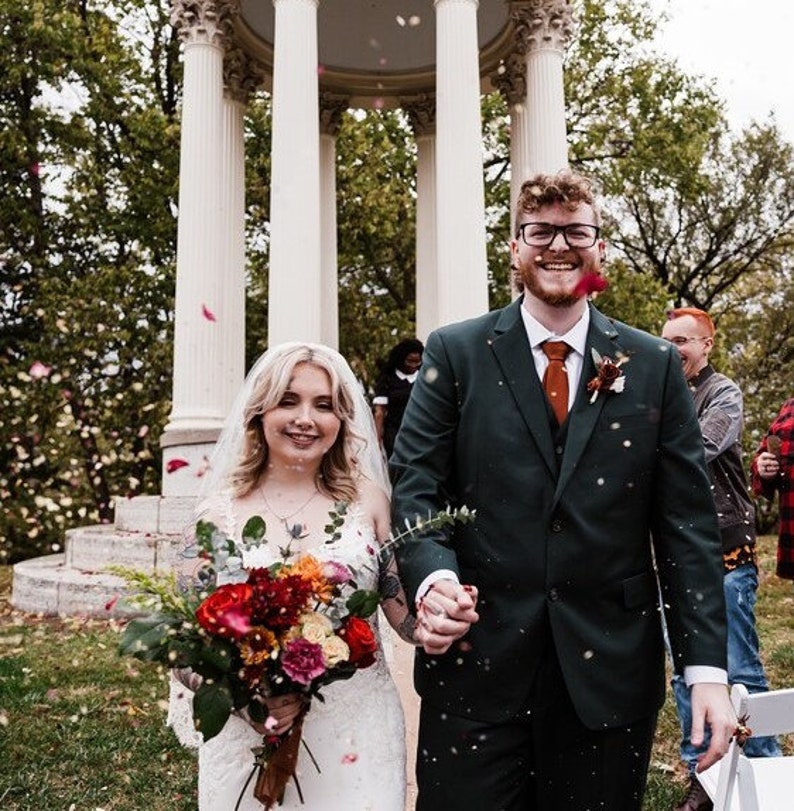 May include: A bride and groom walk down the aisle after their wedding ceremony, with red and white flower petals falling around them. The bride is wearing a white wedding dress with a veil and carrying a bouquet of red, orange, and yellow flowers. The groom is wearing a dark green suit with a red tie. They are both smiling.