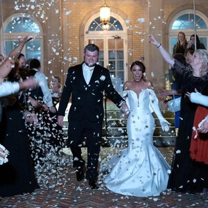 May include: A bride and groom walk down a set of stairs, holding hands, as guests throw white flower petals at them. The bride is wearing a white off-the-shoulder gown with a long train. The groom is wearing a black suit and a white bow tie.