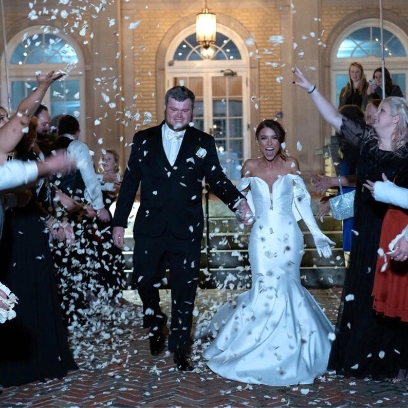 May include: A bride and groom walk down a set of stairs, holding hands, as guests throw white flower petals at them. The bride is wearing a white off-the-shoulder gown with a long train. The groom is wearing a black suit and a white bow tie.