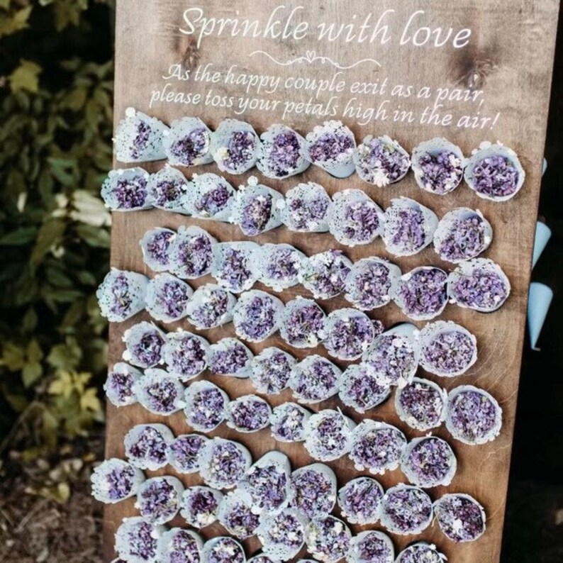 May include: A wooden sign with the text "Sprinkle with love" and "As the happy couple exit as a pair, please toss your petals high in the air!"  The sign is decorated with small, white, lace-edged cones filled with purple flower petals.