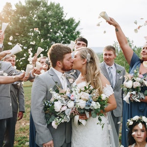 May include: A bride and groom kiss as their wedding party throws flower petals in the air. The bride is wearing a white lace dress and the groom is wearing a gray suit. The wedding party is wearing blue dresses and gray suits. The ceremony is taking place outdoors in a grassy area.