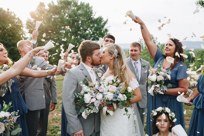 May include: A bride and groom kiss as their wedding party throws flower petals in the air. The bride is wearing a white lace dress and the groom is wearing a gray suit. The wedding party is wearing blue dresses and gray suits. The ceremony is taking place outdoors in a grassy area.
