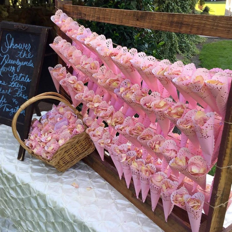 May include: A wooden frame holds pink paper cones filled with rose petals. The cones are arranged in rows and are tied with string. A wicker basket filled with rose petals sits in front of the frame. The sign reads "Shower the Newlyweds with Petals as they start their life as Together".
