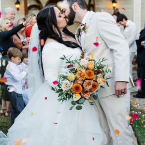 May include: A bride and groom kiss under a shower of rose petals. The bride is wearing a white wedding dress and holding a bouquet of orange, peach, and white flowers. The groom is wearing a white suit.