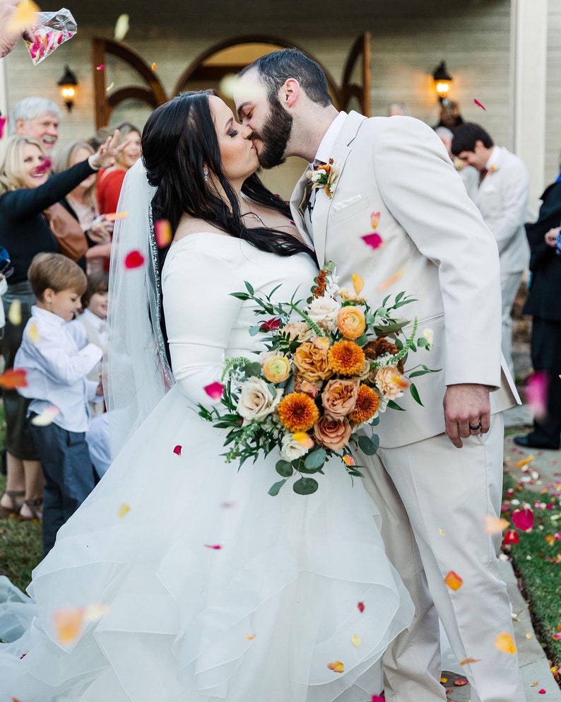 May include: A bride and groom kiss under a shower of rose petals. The bride is wearing a white wedding dress and holding a bouquet of orange, peach, and white flowers. The groom is wearing a white suit.