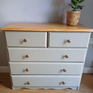 May include: A wooden chest of drawers featuring a natural light-toned wood top and a light grey painted body. The unit has two small top drawers and three full-width lower drawers, each fitted with a round brass-coloured knob handle.