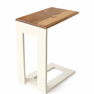 May include: A small, rectangular side table with a two-tone design. The tabletop is made of light and dark brown wood, while the frame is painted white. The table is designed to slide under a sofa or chair.