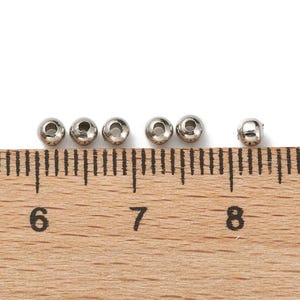 Peut inclure: Six petites perles m&eacute;talliques argent&eacute;es et brillantes avec un trou au centre sont dispos&eacute;es en rang&eacute;e. Une r&egrave;gle en bois avec des graduations en centim&egrave;tres est en dessous, avec les chiffres 6, 7 et 8 visibles.