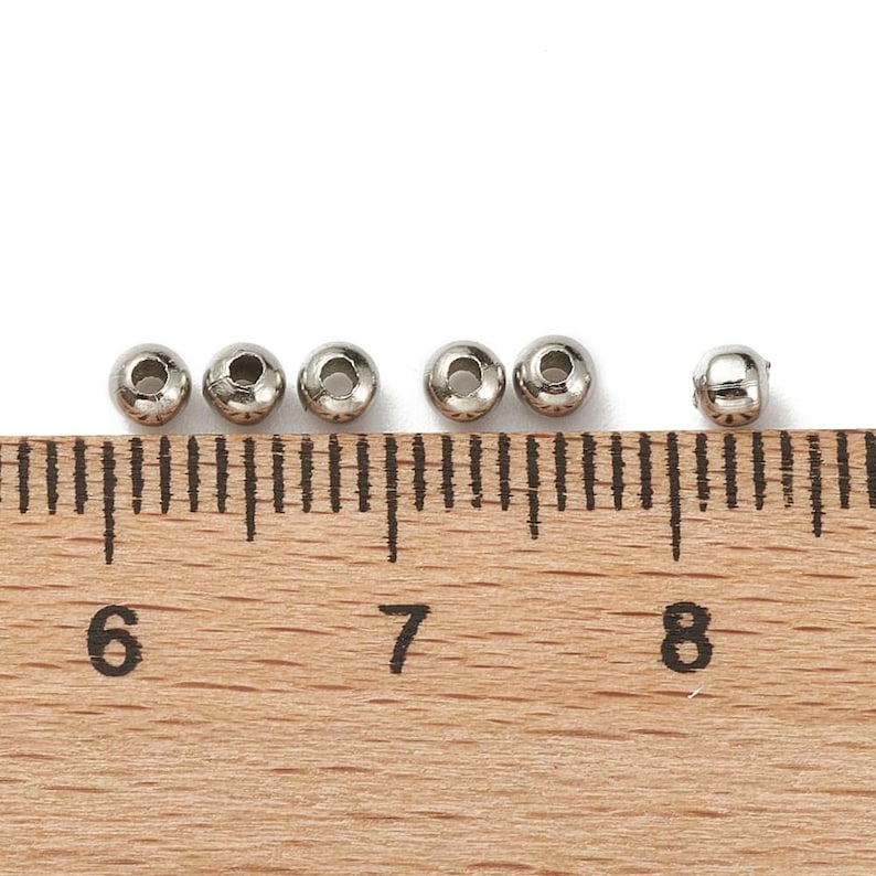 Peut inclure: Six petites perles m&eacute;talliques argent&eacute;es et brillantes avec un trou au centre sont dispos&eacute;es en rang&eacute;e. Une r&egrave;gle en bois avec des graduations en centim&egrave;tres est en dessous, avec les chiffres 6, 7 et 8 visibles.