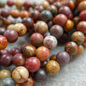 Könnte beinhalten: Nahaufnahme einer Kette aus runden Edelsteinperlen in Erdtönen. Die Perlen sind hauptsächlich in Rot-, Braun-, Gelb- und Grautönen gehalten und weisen einzigartige Muster und Texturen auf. Die Perlen sind aufgefädelt, was auf eine Halskette oder ein Armband hindeutet.