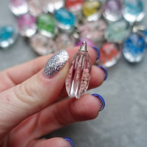 Op de afbeelding: Een heldere, roze, druppelvormige hanger met verticale groeven en een zilverkleurige dop. De hanger wordt vastgehouden in een hand met zilveren glitter nagellak. Andere kleurrijke hangers zijn op de achtergrond.