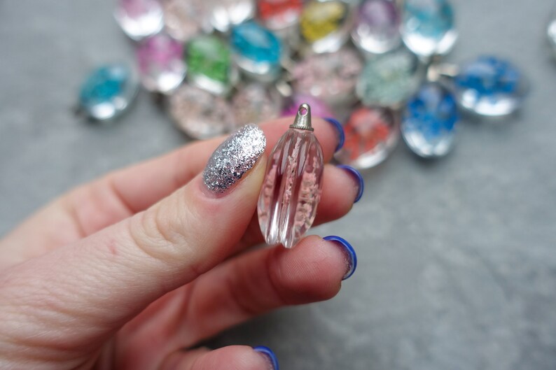 Op de afbeelding: Een heldere, roze, druppelvormige hanger met verticale groeven en een zilverkleurige dop. De hanger wordt vastgehouden in een hand met zilveren glitter nagellak. Andere kleurrijke hangers zijn op de achtergrond.