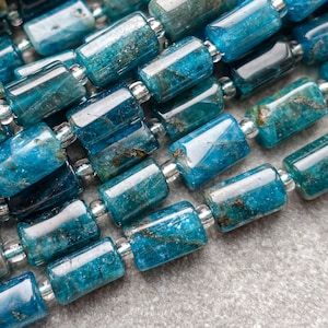 May include: Close-up of strands of blue-green apatite gemstone beads. The beads are cylindrical with a polished finish, separated by small silver-coloured spacers. The colour varies from teal to deep blue, with natural inclusions.