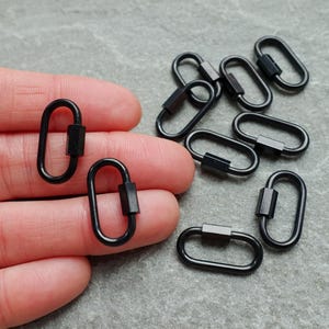 K&ouml;nnte beinhalten: Eine Sammlung kleiner, ovaler Karabinerhaken aus schwarzem Metall. Jeder Karabiner verf&uuml;gt &uuml;ber einen federbelasteten Verschlussmechanismus zum &Ouml;ffnen und Schlie&szlig;en. Das Set enth&auml;lt mehrere einzelne Einheiten, von denen zwei vorne gehalten werden.
