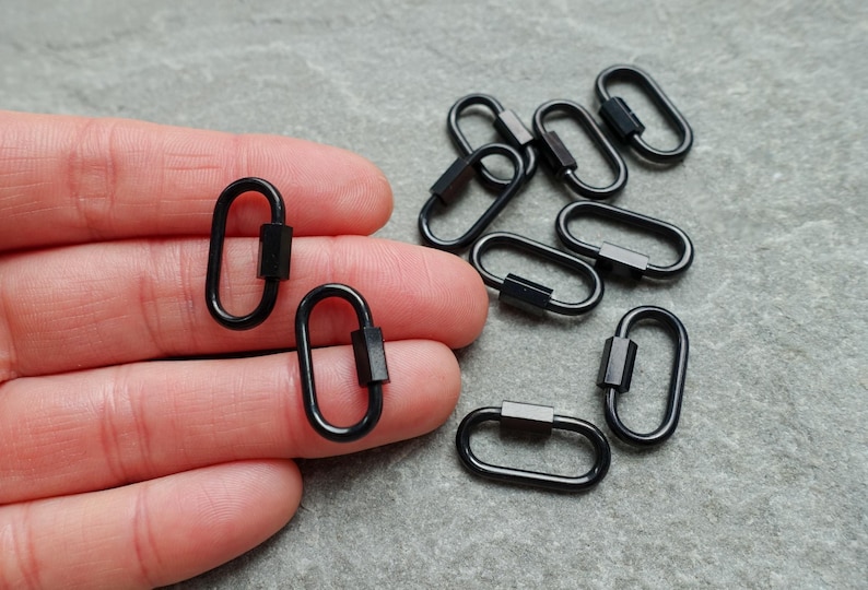 K&ouml;nnte beinhalten: Eine Sammlung kleiner, ovaler Karabinerhaken aus schwarzem Metall. Jeder Karabiner verf&uuml;gt &uuml;ber einen federbelasteten Verschlussmechanismus zum &Ouml;ffnen und Schlie&szlig;en. Das Set enth&auml;lt mehrere einzelne Einheiten, von denen zwei vorne gehalten werden.