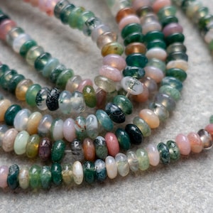 Puede incluir: Primer plano de cuentas de piedras preciosas en tonos verdes, rosas y transparentes. Las pequeñas cuentas planas y redondas están ensartadas. Las cuentas de piedra natural tienen un aspecto único y orgánico, para la creación de joyas.