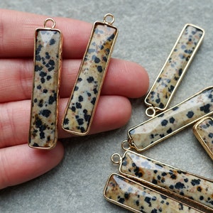Könnte beinhalten: Rechteckige Edelsteinanhänger mit einem gesprenkelten Muster in Beige und Schwarz, gefasst in goldfarbenen Metallrahmen. Jeder Anhänger hat oben eine kleine Schlaufe zum Befestigen an Schmuck. Die Anhänger sind etwa 2,5 cm lang.