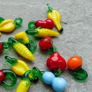 May include: A selection of glass vegetable beads in various colours, such as yellow corn, red tomatoes, and orange peppers. Each bead has a green stem and is arranged on a grey surface. The beads are detailed and appear handmade.