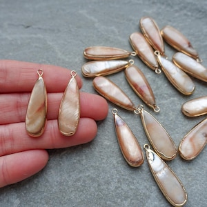 May include: A collection of teardrop-shaped pendants with a pearlescent, beige and brown interior. Each pendant is outlined with a gold-colored metal frame and has a small loop at the top. The pendants are displayed on a gray surface.