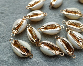 May include: A collection of white and gold cowrie shell charms. Each shell is edged with gold-colored metal and has small loops for attaching to jewelry or other crafts. The shells are arranged on a gray surface.