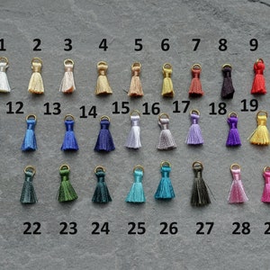 May include: A collection of 29 small tassels in various colors, each with a gold-colored loop at the top. The tassels are arranged in rows, showcasing a wide spectrum of hues, from white to black, and including shades of blue, green, and pink.