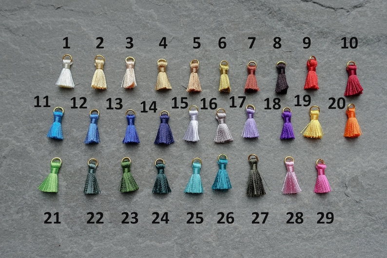 May include: A collection of 29 small tassels in various colors, each with a gold-colored loop at the top. The tassels are arranged in rows, showcasing a wide spectrum of hues, from white to black, and including shades of blue, green, and pink.