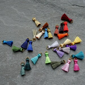 May include: A collection of small, colorful tassels with gold-colored metal loops. The tassels are in various colors, including blue, green, purple, pink, and yellow. They are scattered on a dark gray surface.
