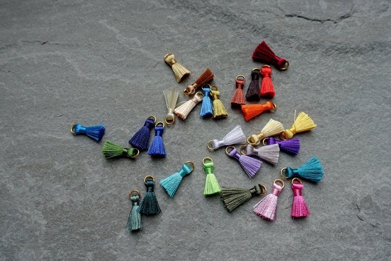 May include: A collection of small, colorful tassels with gold-colored metal loops. The tassels are in various colors, including blue, green, purple, pink, and yellow. They are scattered on a dark gray surface.