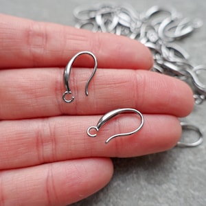 May include: Close-up of several silver-colored earring hooks. Two hooks are in focus, showing a curved design with a small loop at the bottom. The hooks are on a hand, with more hooks visible in the background.