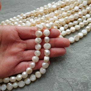 Könnte beinhalten: Nahaufnahme von Strängen cremeweißer, unregelmäßig geformter Süßwasserperlen. Die Perlen sind aufgereiht. Die Perlen werden in einer Hand gehalten, mit einem grauen, strukturierten Hintergrund.