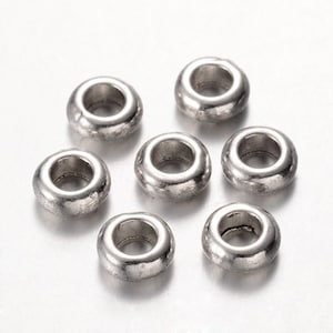 Peut inclure: Sept perles rondes argentées avec un trou au centre sont disposées sur un fond clair. Les perles ont une surface légèrement texturée et un éclat métallique, suggérant qu'elles sont en métal.