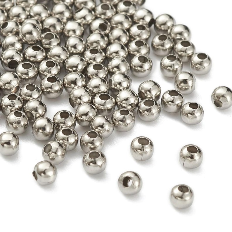 Peut inclure: Un ensemble de perles m&eacute;talliques argent&eacute;es et brillantes. Chaque perle est ronde avec un trou traversant le centre. Les perles sont dispers&eacute;es sur une surface blanche, refl&eacute;tant la lumi&egrave;re.