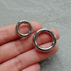 Könnte beinhalten: Zwei glänzende, silberfarbene Metall-Federringe in verschiedenen Größen. Die Ringe sind kreisförmig mit einer Scharnieröffnung. Ein Ring ist kleiner als der andere. Die Ringe werden auf einer Hand gezeigt.